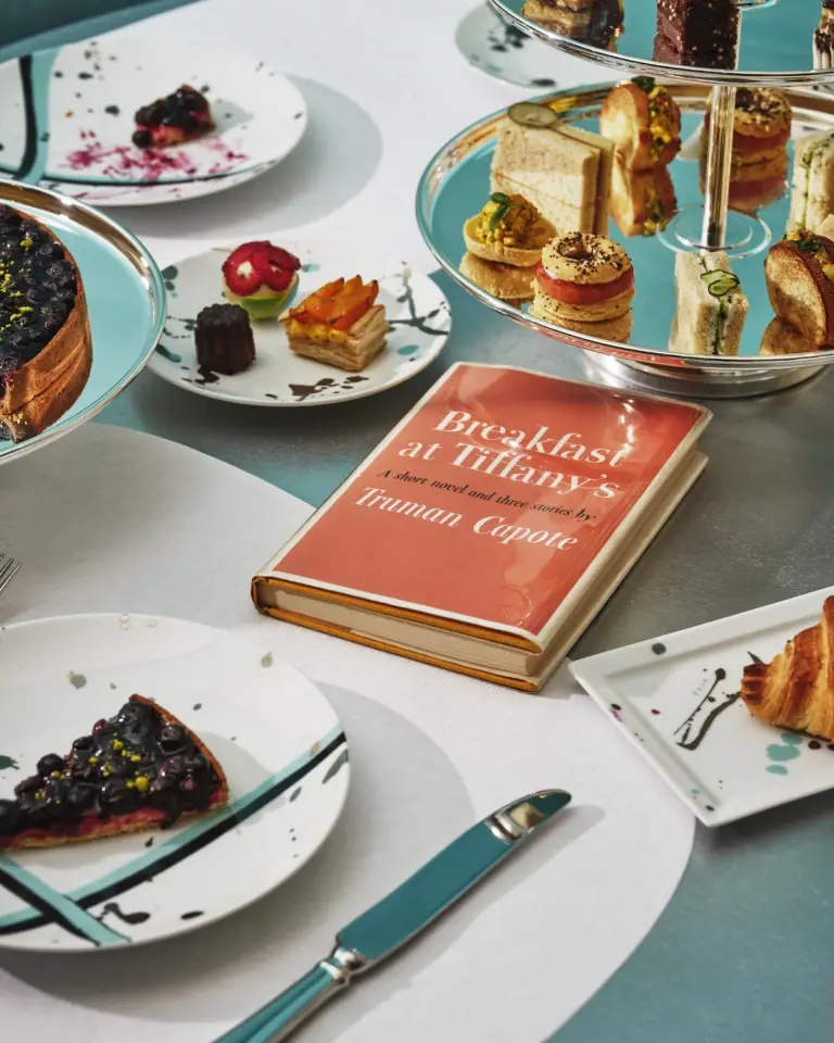An elegant breakfast spread with pastries, cakes, and a tiered stand of sandwiches is arranged on a table, with a physical…