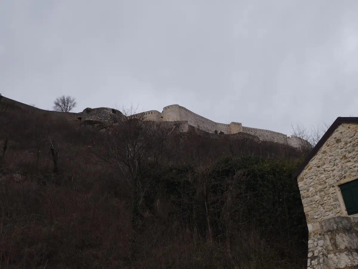 A photograph shows a long, stone castle wall built along a hillside under an overcast sky, with bare trees and shrubs in the…