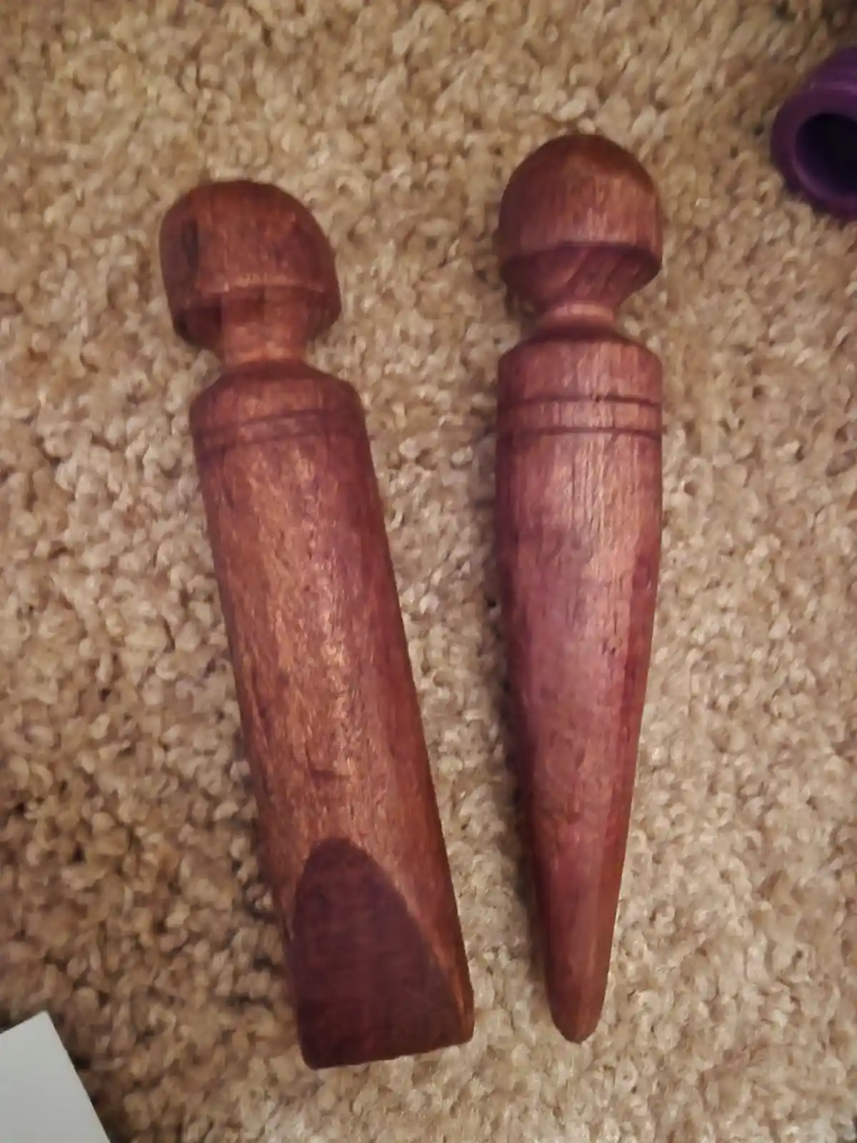 Two hand-carved wooden dildos lie side by side on a beige carpet, one with a blunt, rectangular tip and the other with a…