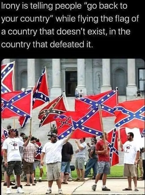 A group of people are gathered outdoors in front of a large classical-style building, holding numerous Confederate battle…