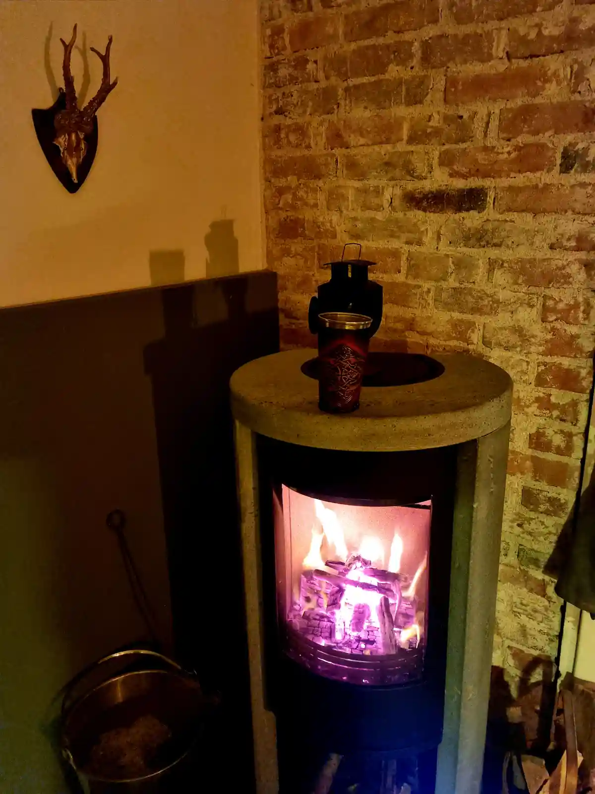 A cozy indoor scene featuring a lit wood-burning stove with glowing flames, a decorative cup and lantern on top, a brick…