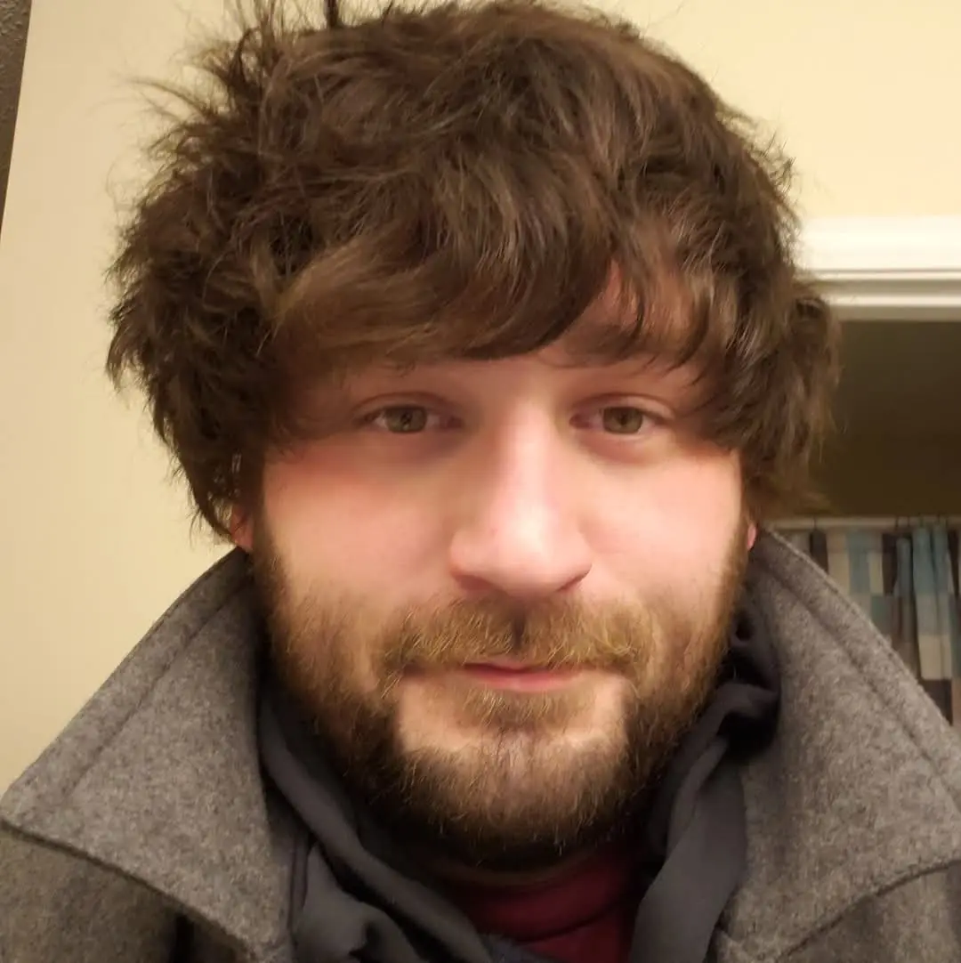 A close-up photo of a man with messy brown hair and a beard, wearing a grey coat over a dark hoodie and red shirt, looking…