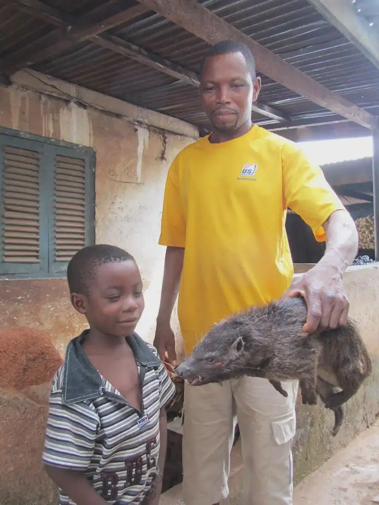 A man in a yellow shirt holds a small, furry, wild-looking animal with a long snout and sharp teeth, while a young boy in a…