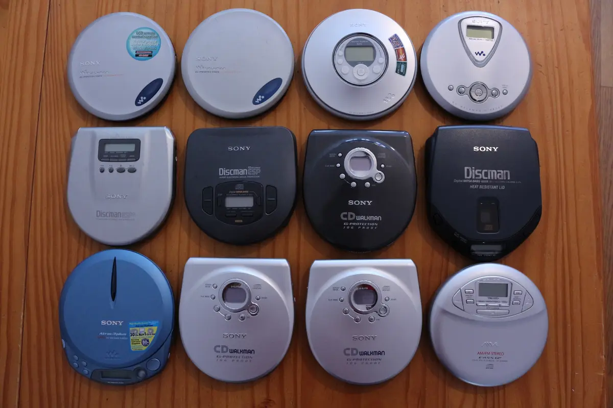 Ten Sony portable CD players of various models and colors are arranged in three rows on a wooden surface. The overlaid text…