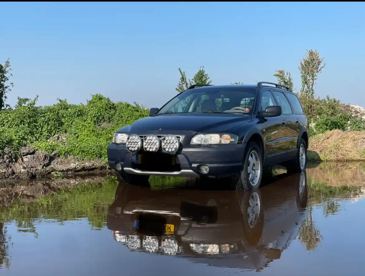 A dark blue station wagon with three large auxiliary lights mounted on its front bumper is parked in a shallow, muddy…