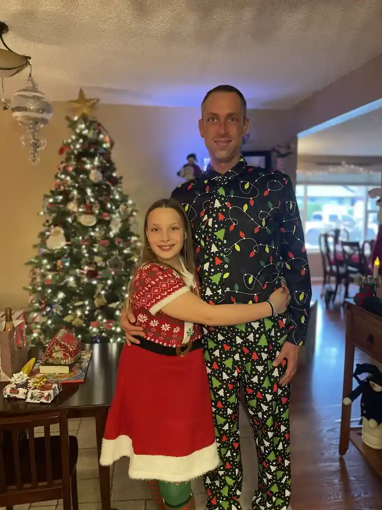 A man and a young girl pose together in a festively decorated living room with a lit Christmas tree in the background. The…