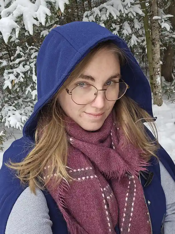 A young woman with blonde hair and glasses wears a blue hooded jacket, a maroon scarf, and a grey shirt, standing in a snowy…