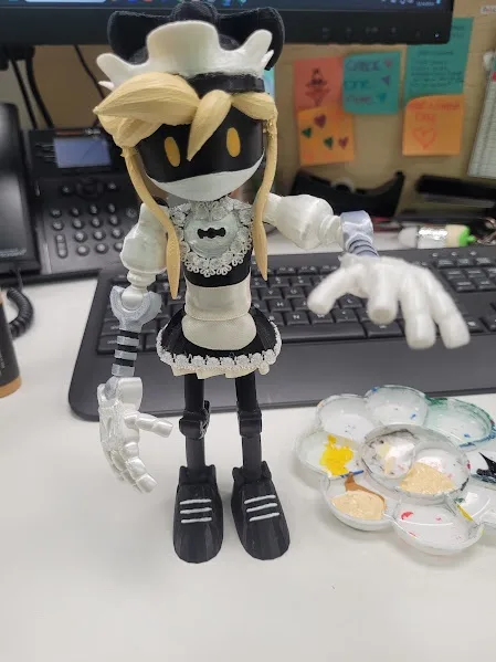 A custom-made doll resembling the character 'Maiden' from the game 'Undertale' is posed on a desk next to a paint palette…