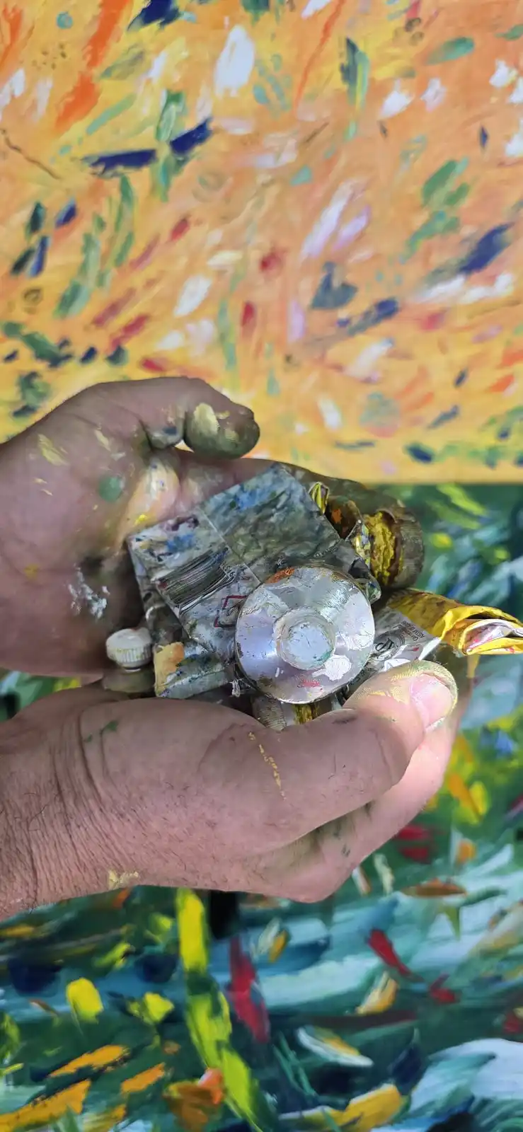 A pair of paint-splattered hands holds a crumpled, used CD and other art supplies against a colorful abstract painting…