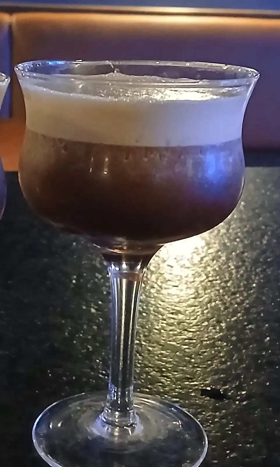 A close-up photo of a cocktail in a stemmed glass, featuring a dark brown liquid base topped with a thick, creamy white…
