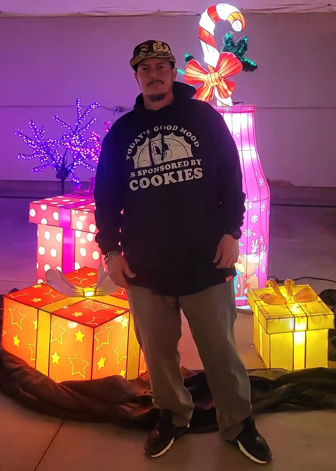A man stands in front of large, illuminated Christmas-themed decorations including gift boxes, a candy cane, and a purple…