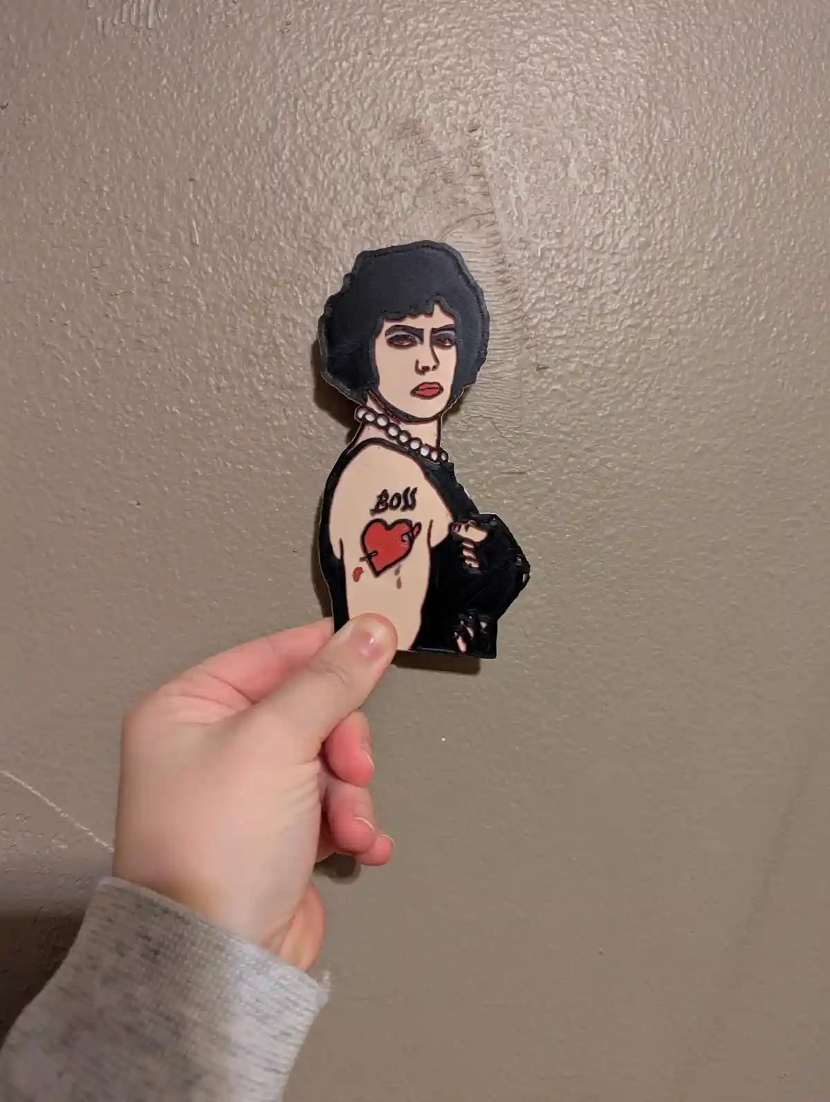 A hand holds a 3D-printed figurine of a woman with a black bob haircut, pearl necklace, and torn black top, showing a tattoo…
