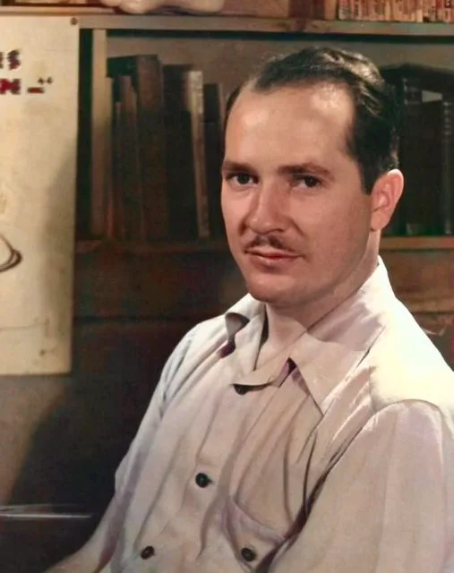 A vintage photo shows a man with a mustache wearing a light-colored button-up shirt, seated in front of a bookshelf. The…