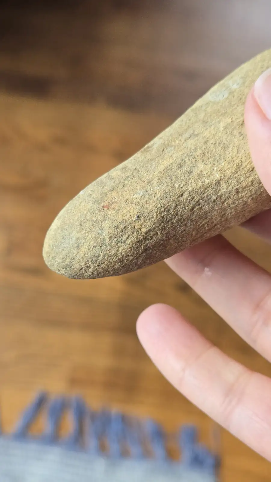 A close-up photo shows a hand holding a smooth, elongated, tan-colored rock with a pointed tip, against a blurred wooden…