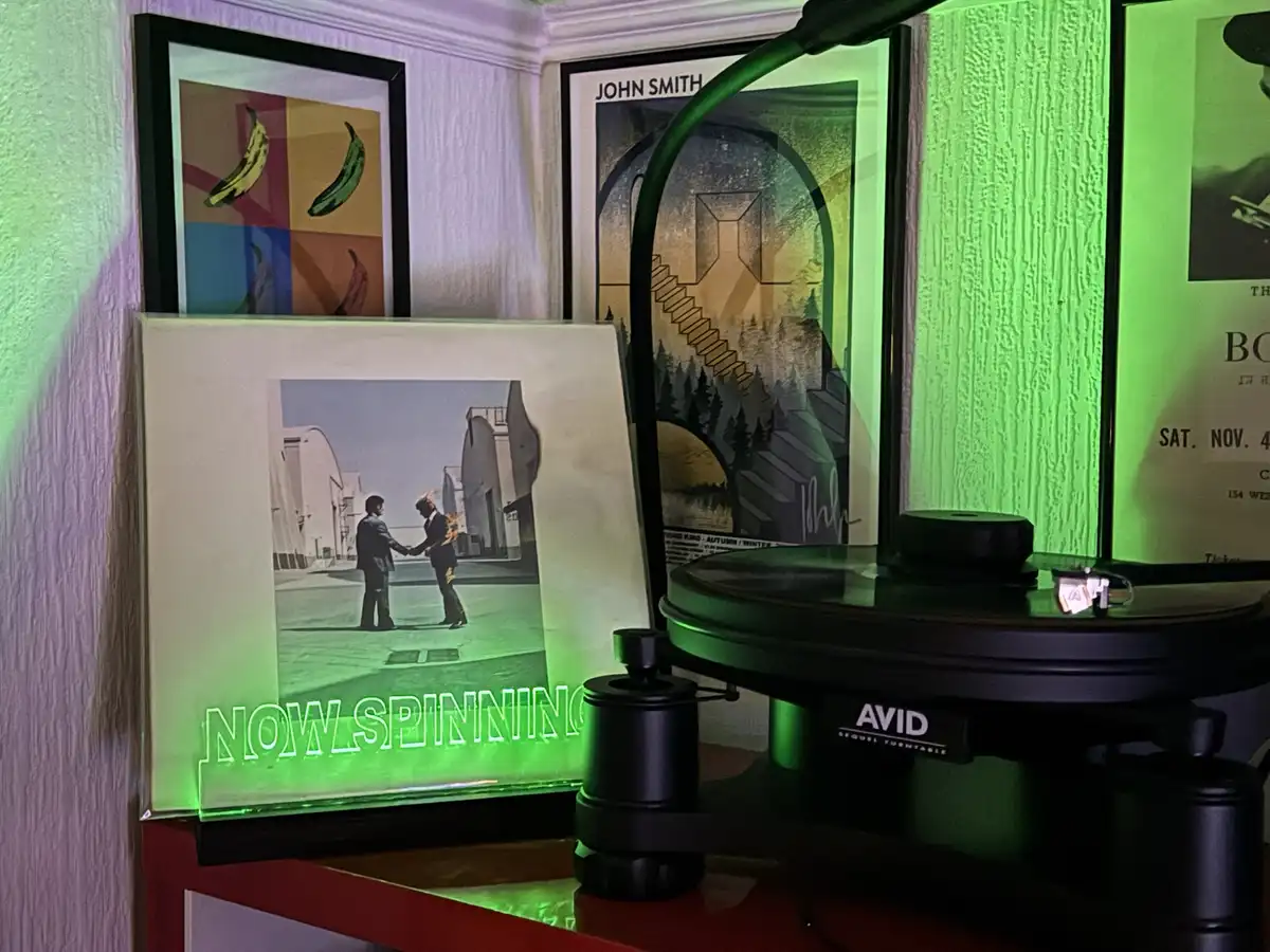 A vinyl record player with a glowing green 'NOW SPINNING' sign sits on a red shelf, next to framed art including a pop-art…