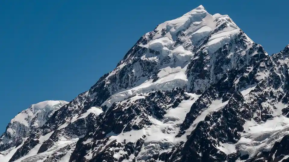 A majestic, snow-capped mountain peak with rugged, dark rock faces under a clear blue sky. The image has white text overlaid…