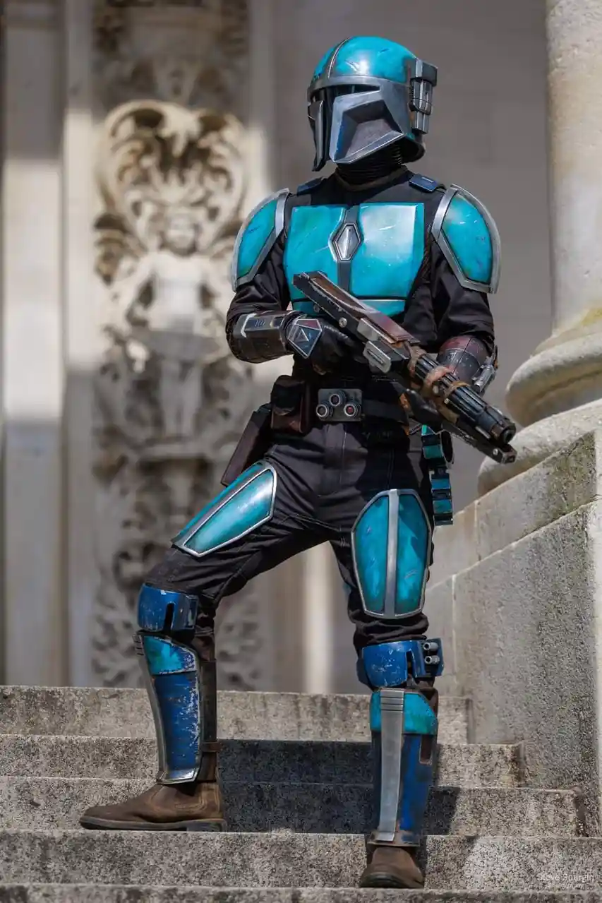 A person in a detailed, weathered blue and black Mandalorian armor costume stands confidently on stone steps, holding a…