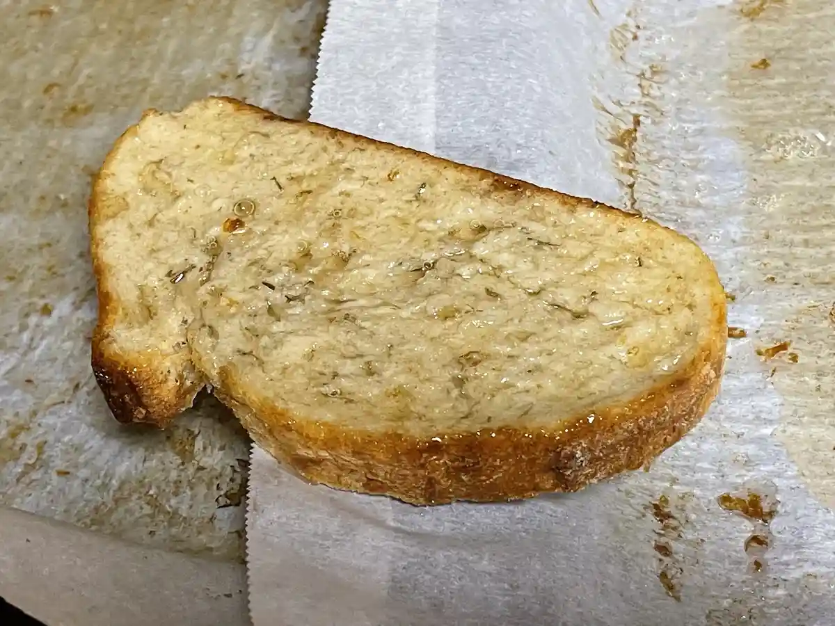 A close-up photo shows a single slice of toasted bread, glistening with oil or butter and sprinkled with herbs, resting on a…
