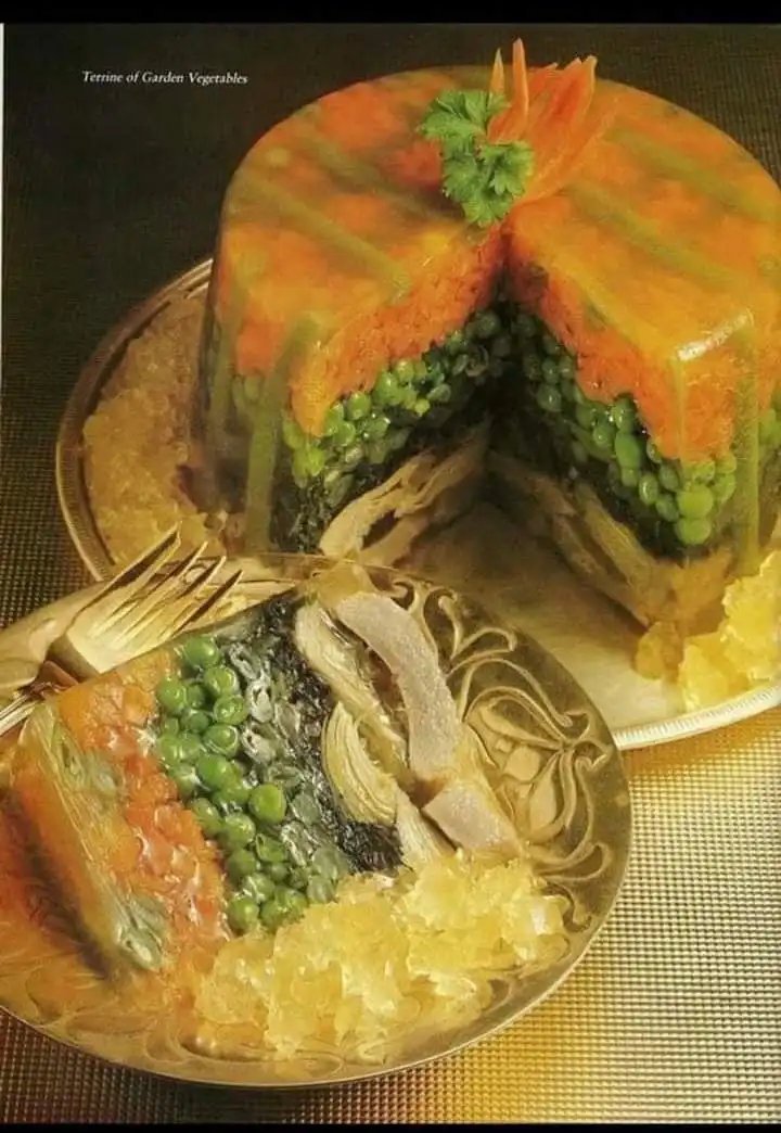 A vintage cookbook photo shows a layered vegetable terrine with a slice removed and served on an ornate plate, revealing…