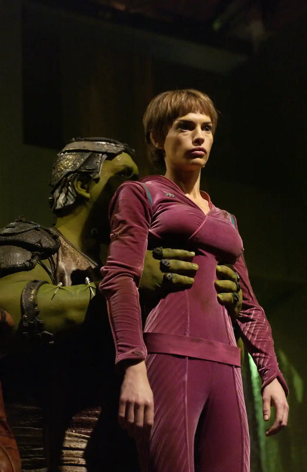 A woman in a purple jumpsuit stands with a stern, unimpressed expression as a green-skinned alien with armor places both…