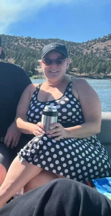 A woman in a black and white polka dot dress and sunglasses sits on a boat, holding a silver thermos. Behind her is a scenic…