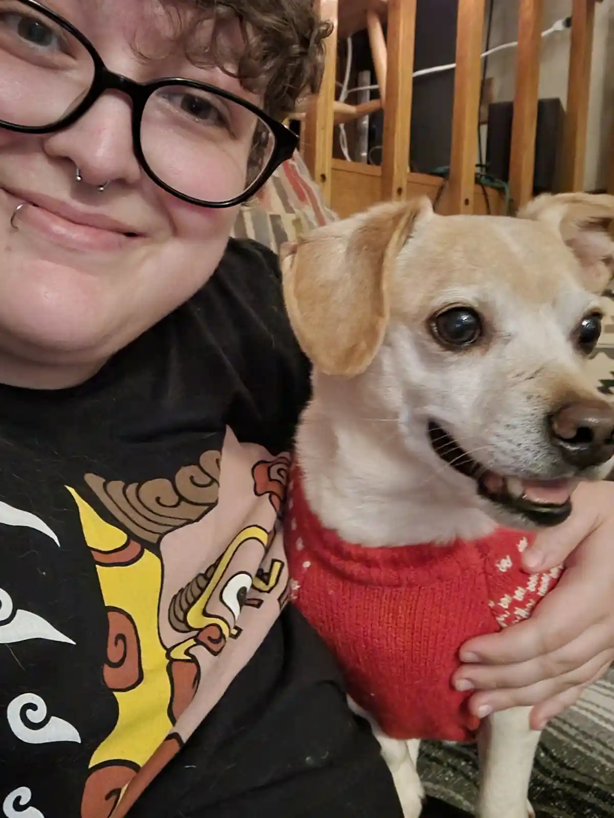 A person with curly hair, black-rimmed glasses, and facial piercings smiles while holding a small, light-colored dog wearing…