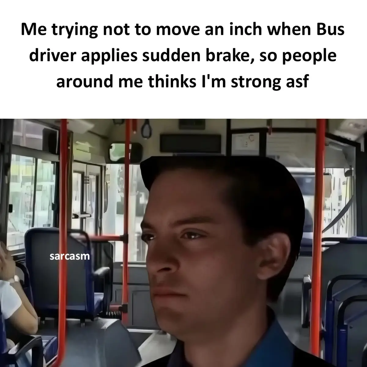 A close-up of a man with a deadpan, unimpressed expression sitting on a bus, with red poles and windows visible in the…
