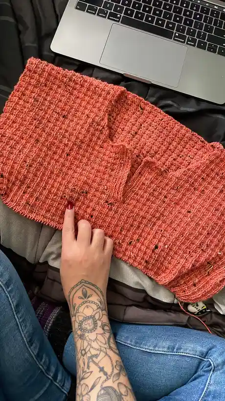 A person with a tattooed arm and red nail polish points at a large, orange, knitted scarf with a ribbed pattern, which has…