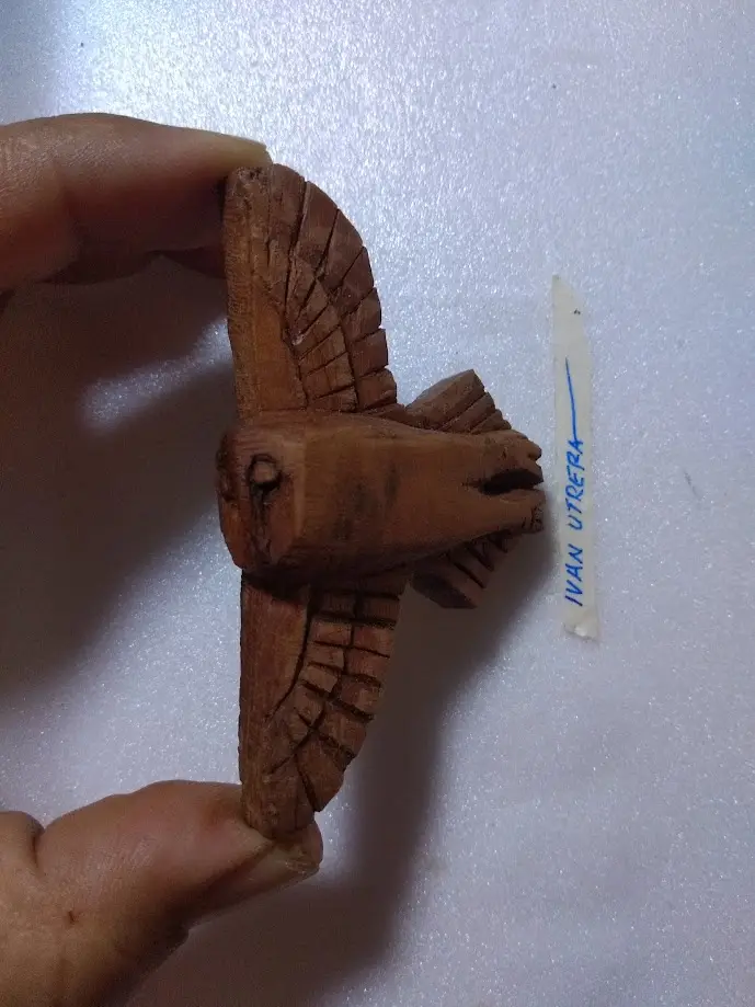 A hand holds a small, crudely carved wooden bird with spread wings against a white textured background. Beside it, a white…