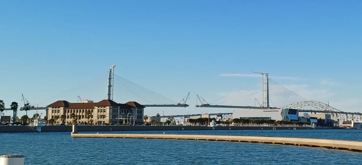 A scenic waterfront photo shows a large cable-stayed bridge under construction with cranes visible, alongside a building and…
