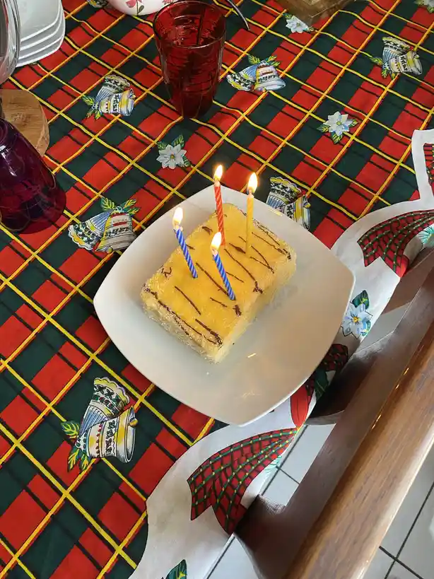 A rectangular yellow cake with chocolate drizzle sits on a white plate atop a festive red, green, and yellow plaid…