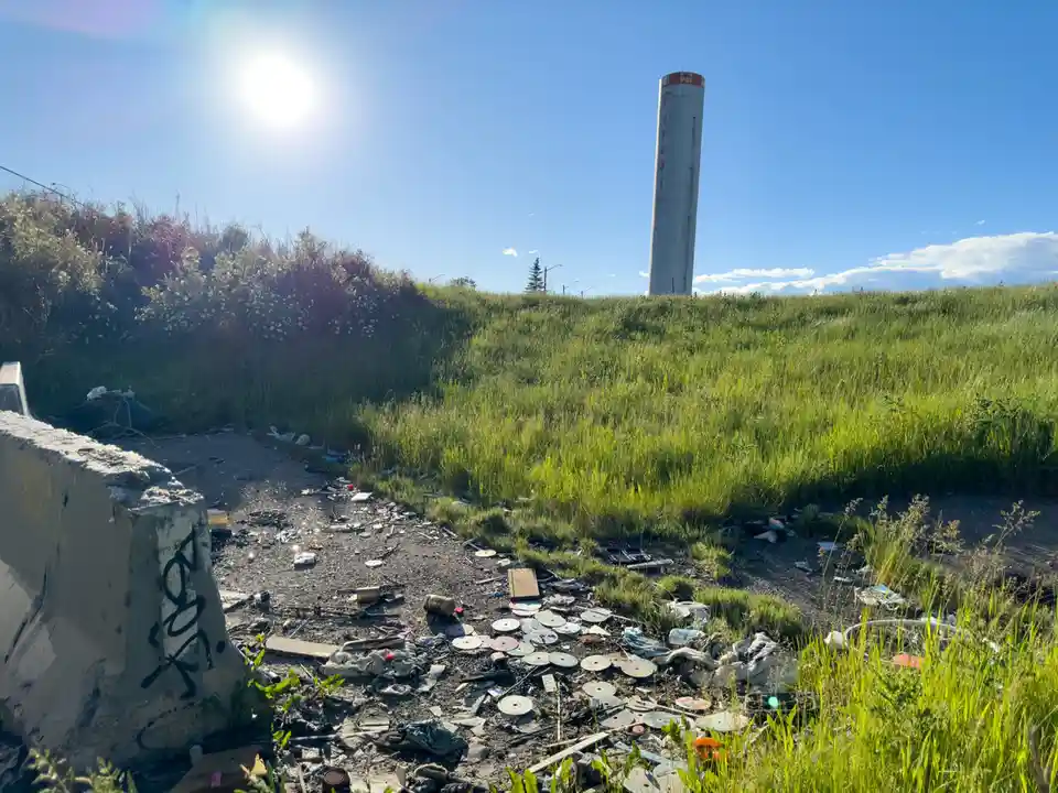 A bright, sunny day shows a grassy field littered with trash, including many broken CDs and discarded items, with a tall…