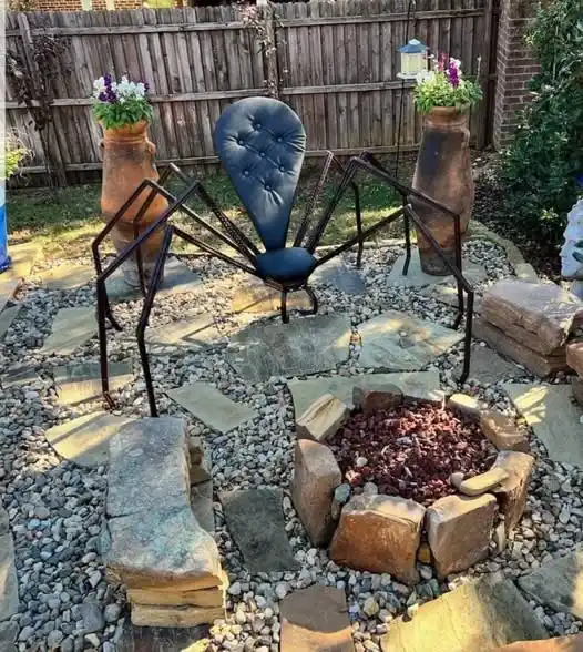 A large, black metal spider-shaped chair with a tufted blue cushion sits in a backyard garden, positioned near a stone fire…