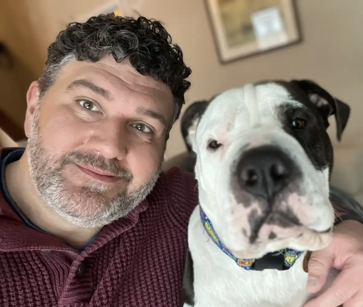 A man with curly dark hair and a graying beard, wearing a maroon textured shirt, takes a selfie with a large white and black…