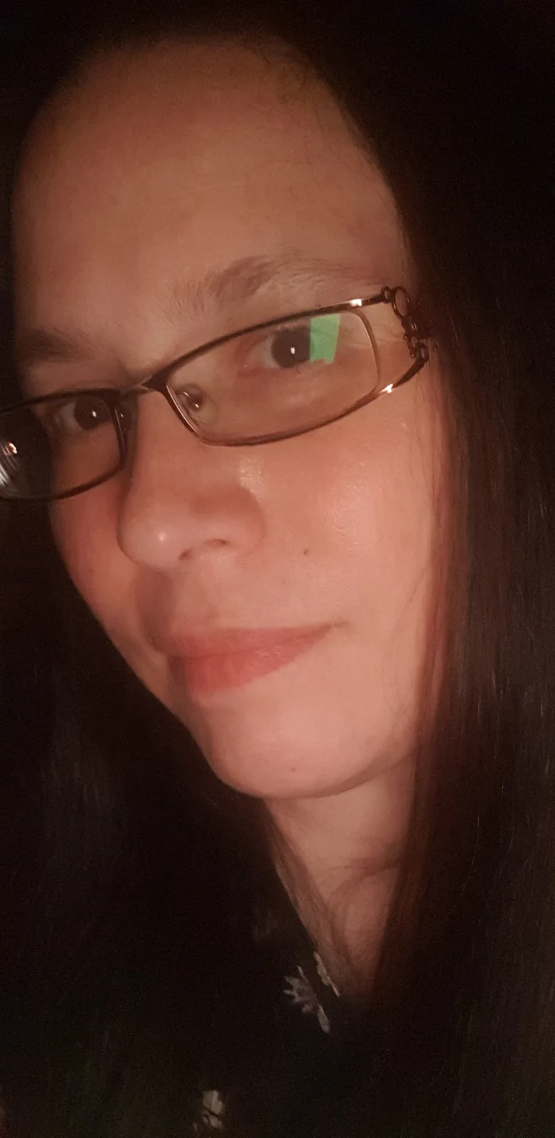 A close-up selfie of a person with dark hair and glasses, looking slightly to the side with a subtle smirk. The lighting is…
