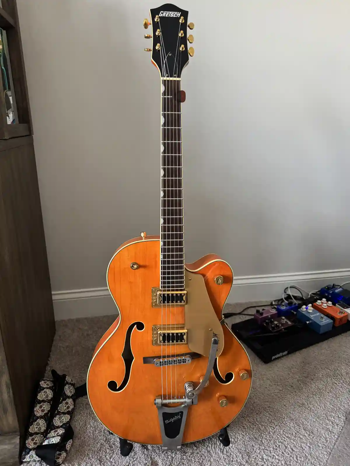 A sunburst-finish Gretsch electric guitar with a Bigsby vibrato tailpiece stands upright on a carpeted floor, next to a…