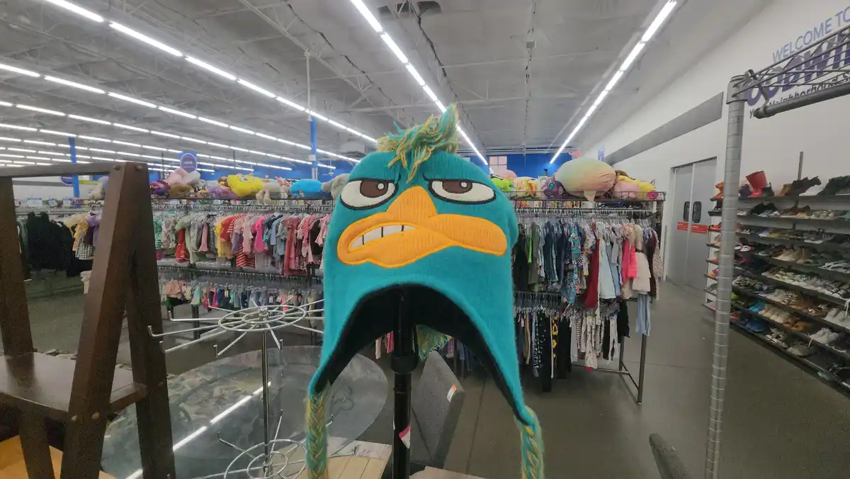 A teal knitted hat featuring the angry face of the animated character Perry the Platypus is displayed on a stand in a…