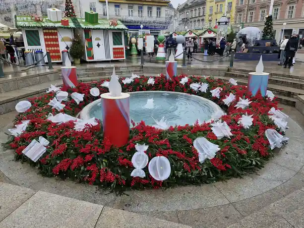 A festive outdoor fountain surrounded by a large wreath of greenery, red berries, and white decorative elements like…