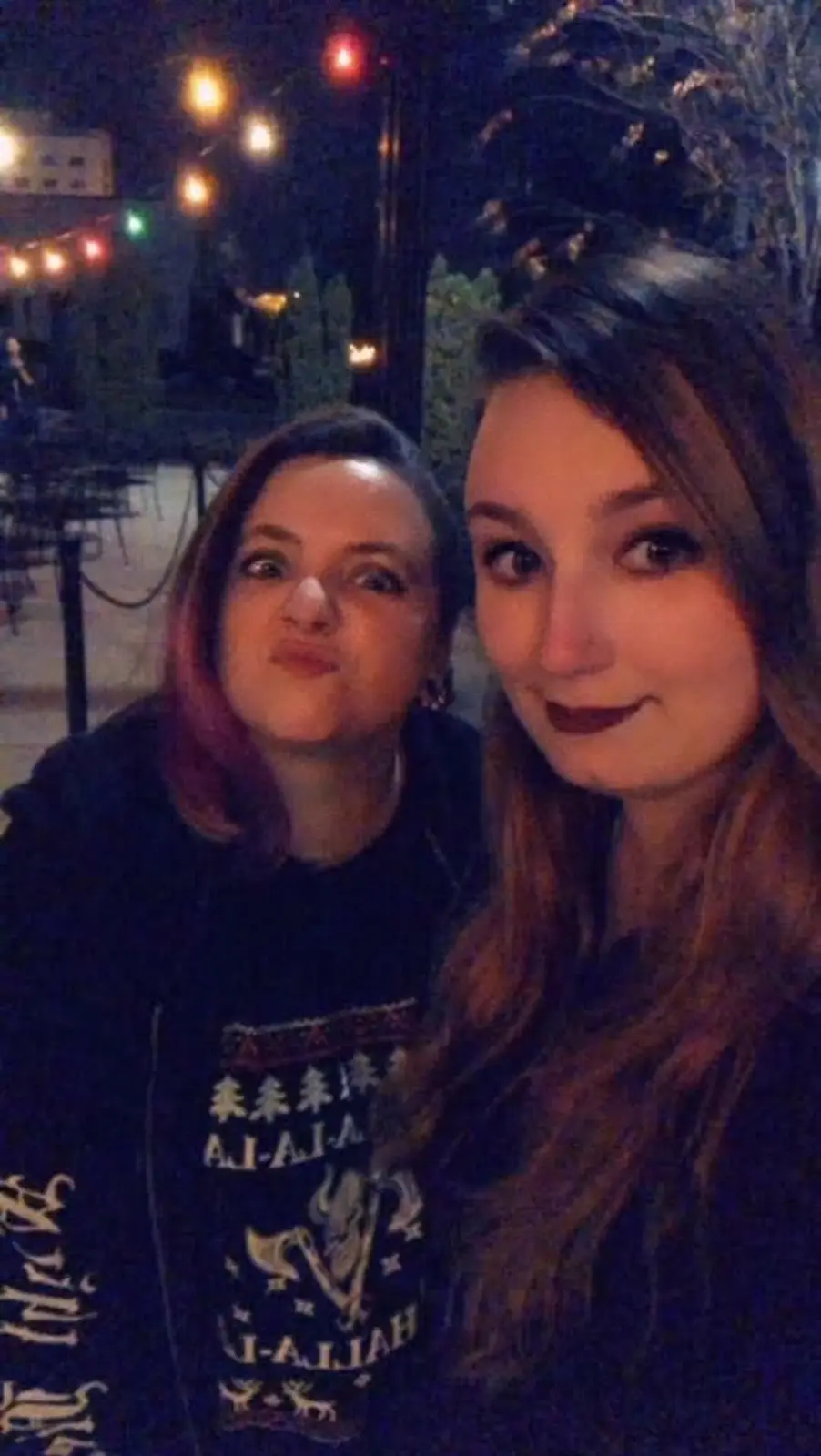 Two women are taking a selfie at night outdoors under string lights. The woman on the left, wearing a dark sweater with a…