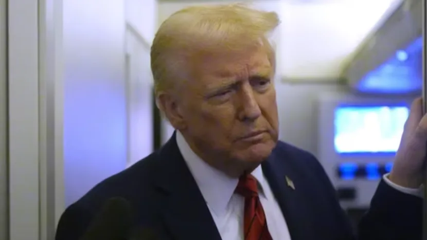 a man with blond hair, wearing a dark suit and red tie, looks concerned while standing in what appears to be an airplane…
