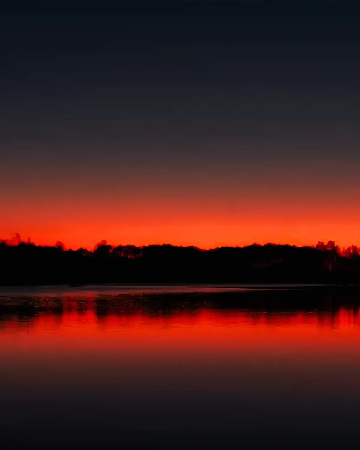 A blurry, motion-blurred photograph of a fiery orange sunset over a dark silhouette of hills or trees, reflected in water…