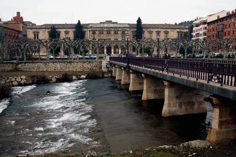 A wide shot of a river flowing under a stone bridge with ornate railings, leading to a large classical building labeled…