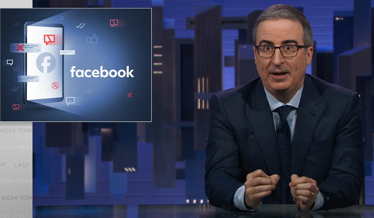 A man in a suit and glasses, likely John Oliver, sits at a desk with a shocked expression, gesturing with his hands. To his…