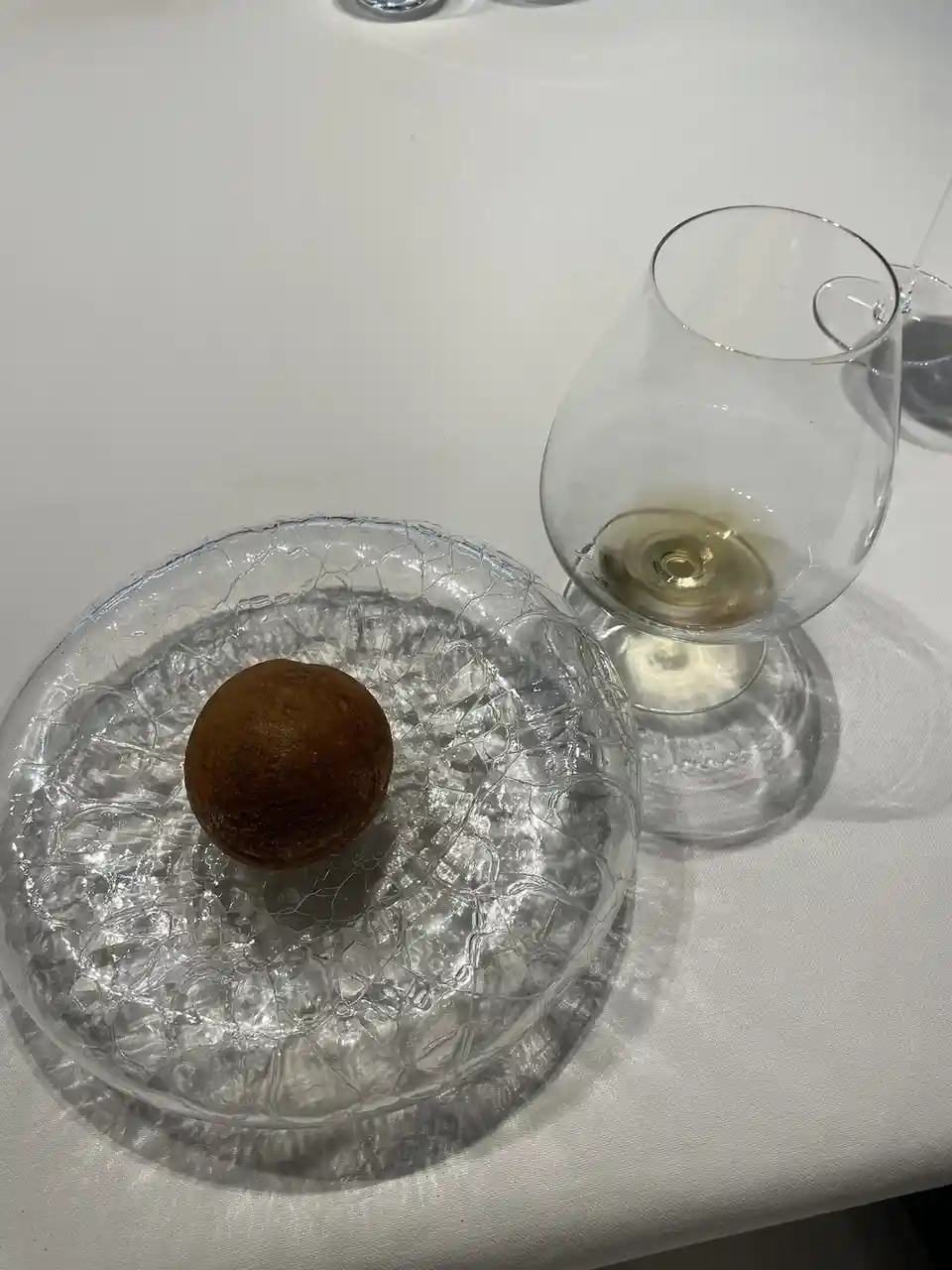 A close-up photo shows a single, small, round, brown dessert resting on a clear, textured glass plate on a white tablecloth…