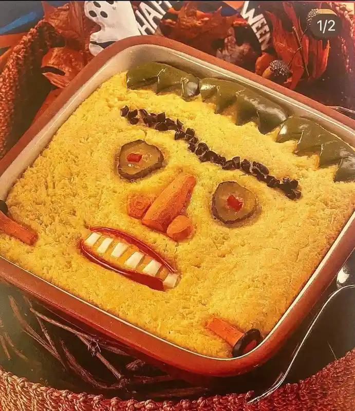 A square pan of yellow cornbread is decorated to look like a scary Halloween face, using green bell pepper strips for hair…
