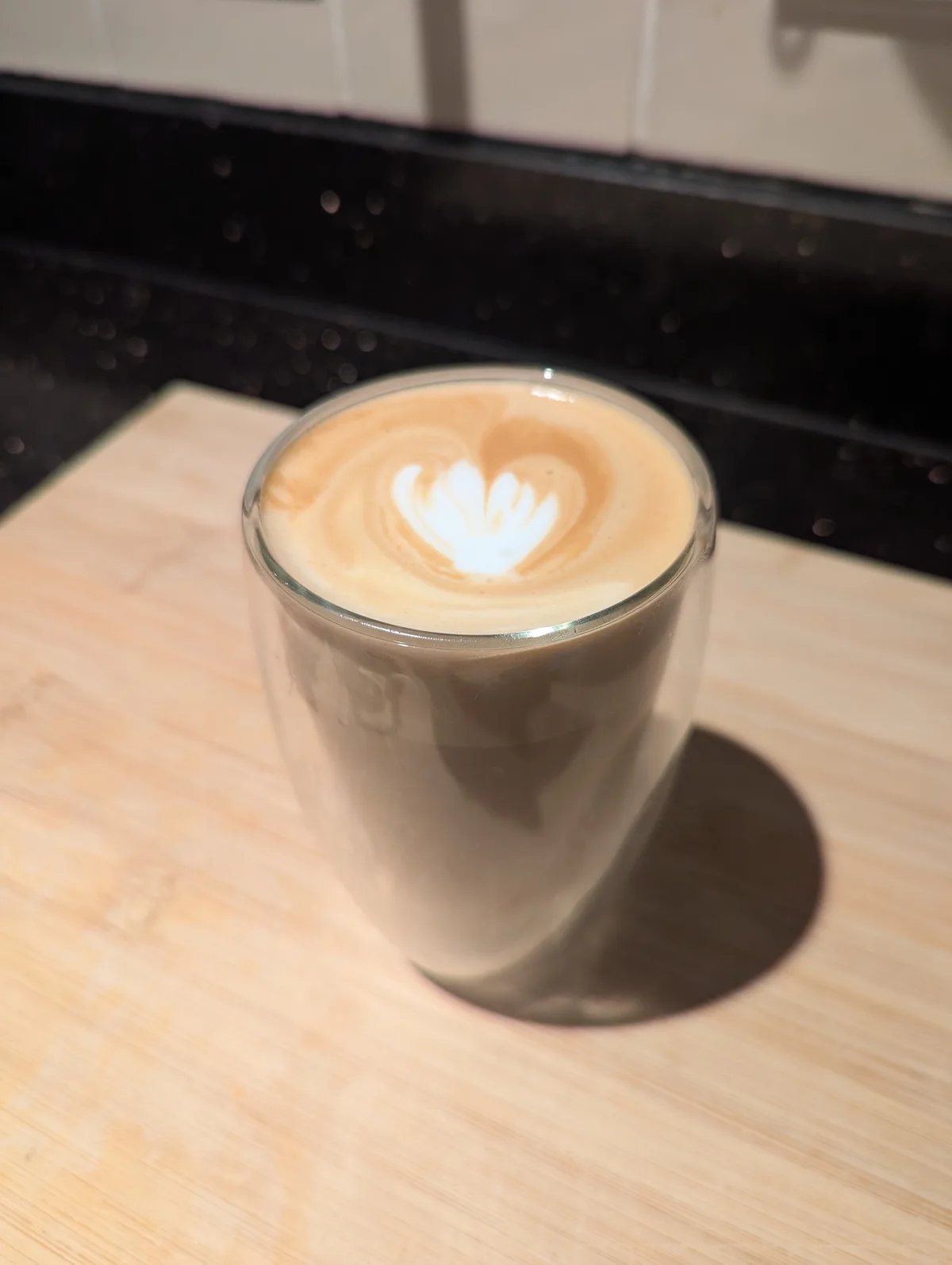 A clear glass cup of coffee with latte art shaped like a heart sits on a light wooden cutting board, with a dark speckled…