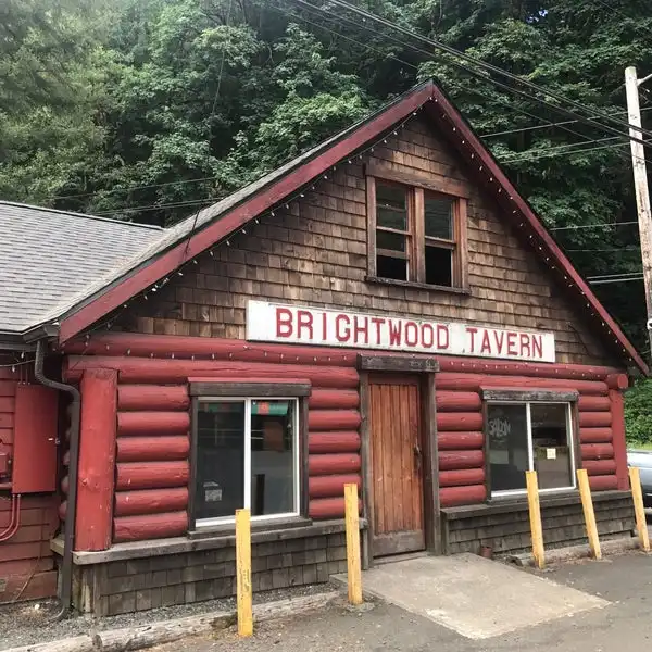 A red-painted log cabin-style building with a sign reading 'BRIGHTWOOD TAVERN' above its wooden door. The structure has…