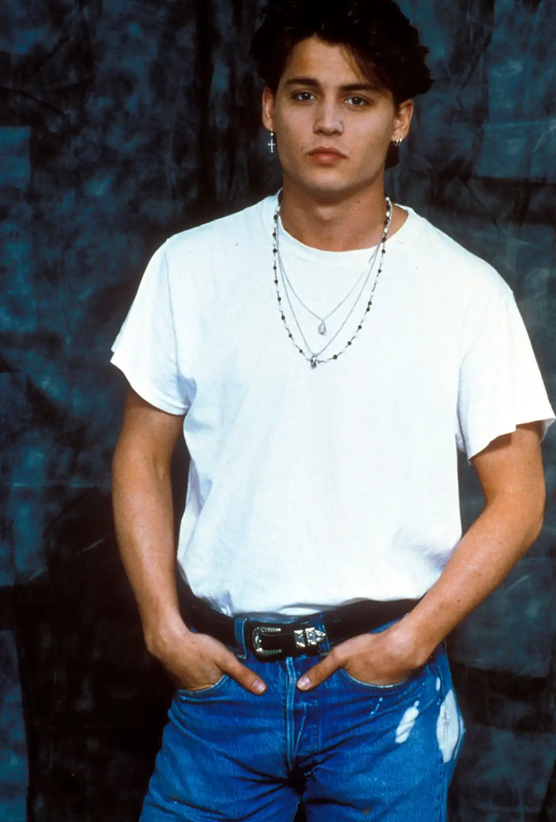 A young man with dark hair and multiple necklaces stands against a blue backdrop, wearing a white t-shirt, blue jeans, and a…