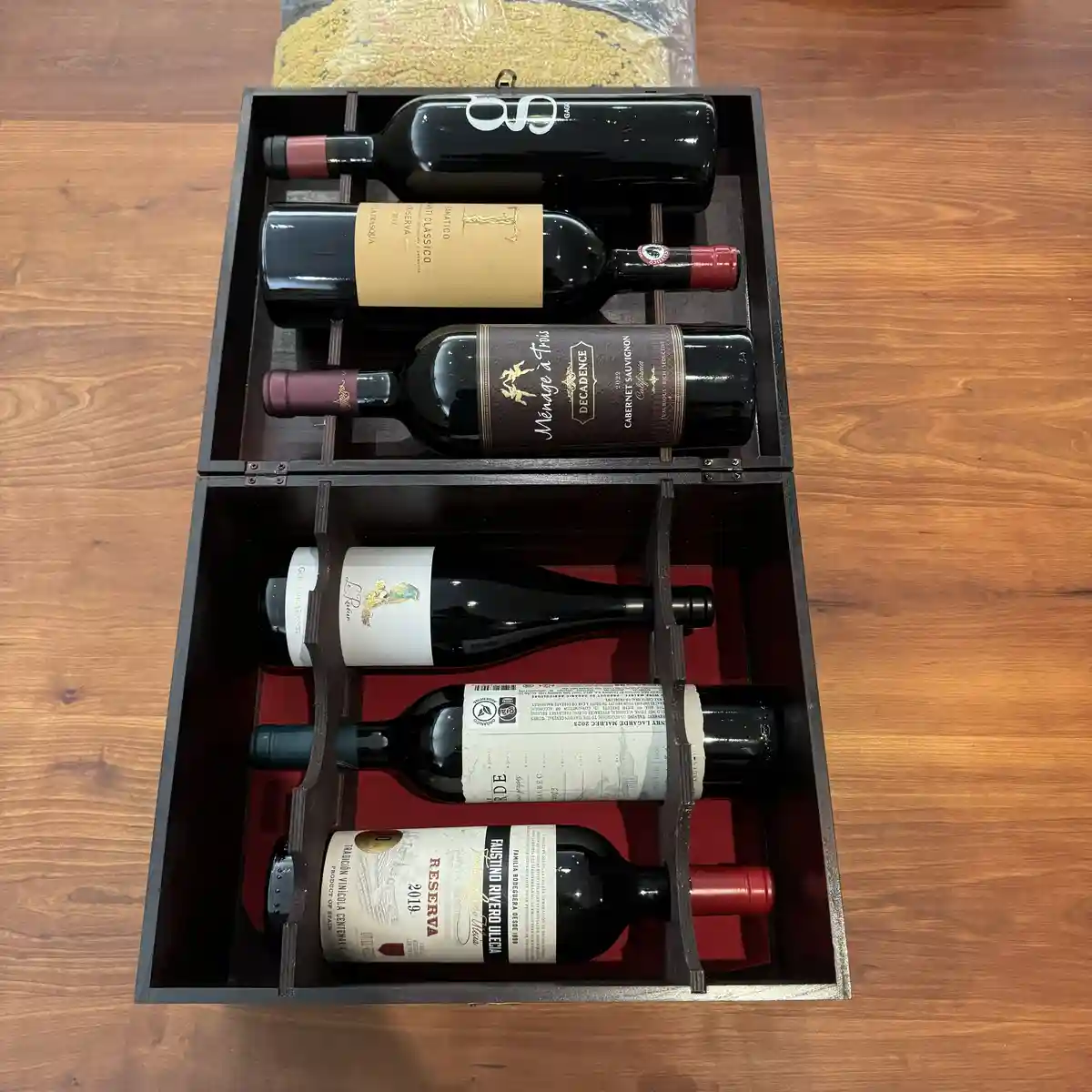 An overhead shot shows a dark wooden wine box open on a wooden floor, revealing six wine bottles neatly arranged in two…