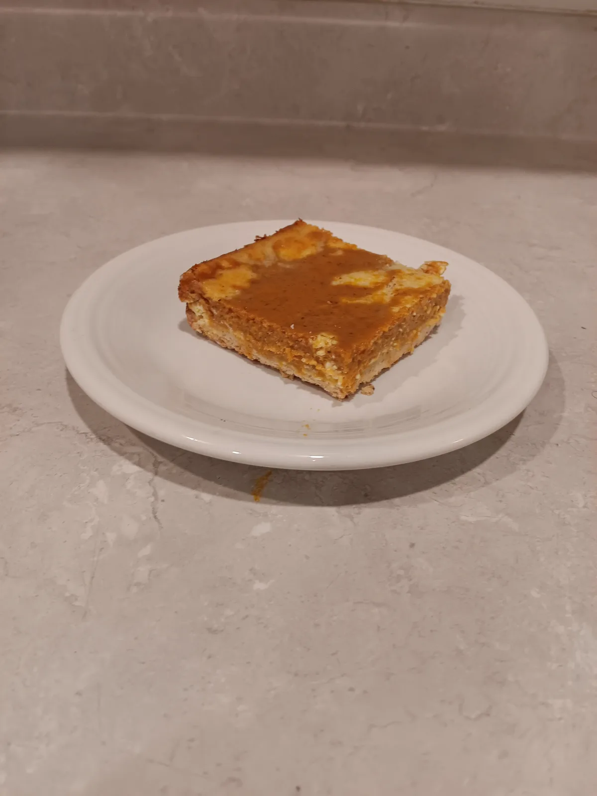 A single square pumpkin pie bar sits on a white plate atop a light-colored countertop. The bar has a golden-brown crust and…